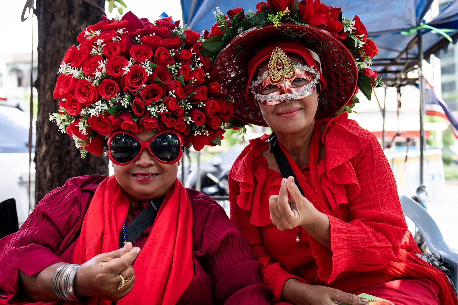 Two women wearing red gesture with their hands. One is in a hat of red roses, the other is wearing elaborate eyewear.